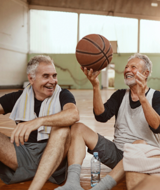 Two senior men sitting on a gymnasium floor while one holds a basketball